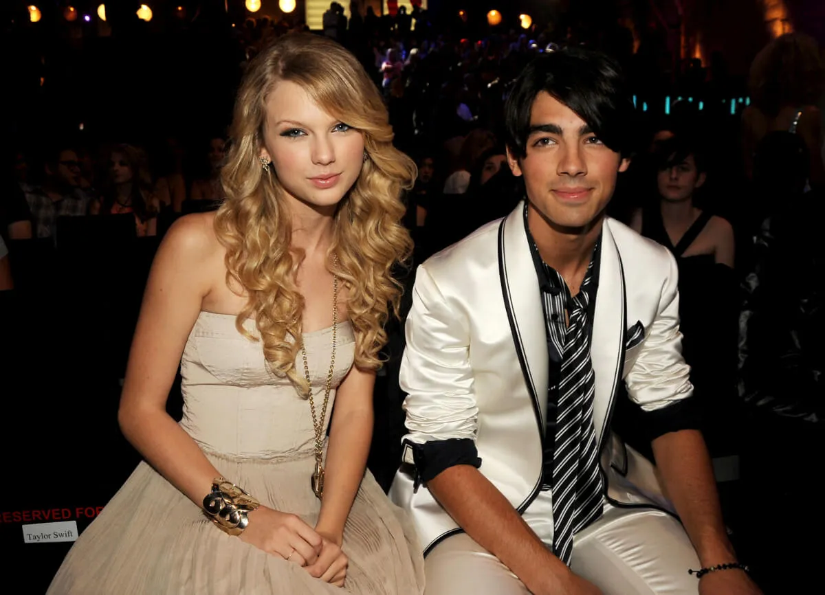 Taylor Swift porte une robe beige et est assise à côté de Joe Jonas, qui porte une veste blanche.