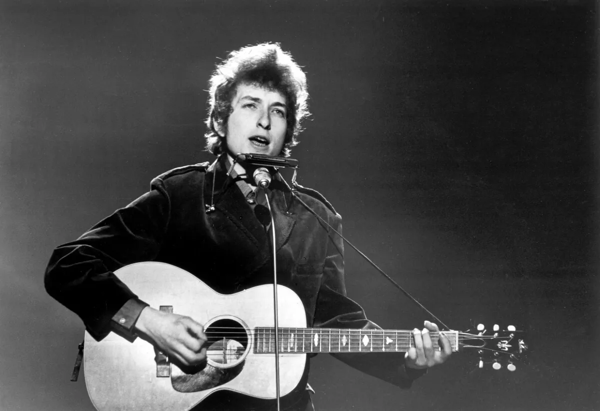 Une photo en noir et blanc de Bob Dylan jouant de la guitare et chantant dans un microphone.