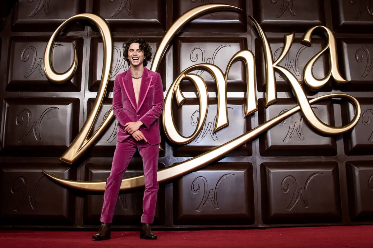 Timothée Chalamet porte un costume rose et se tient devant une barre de chocolat pour « Wonka ».