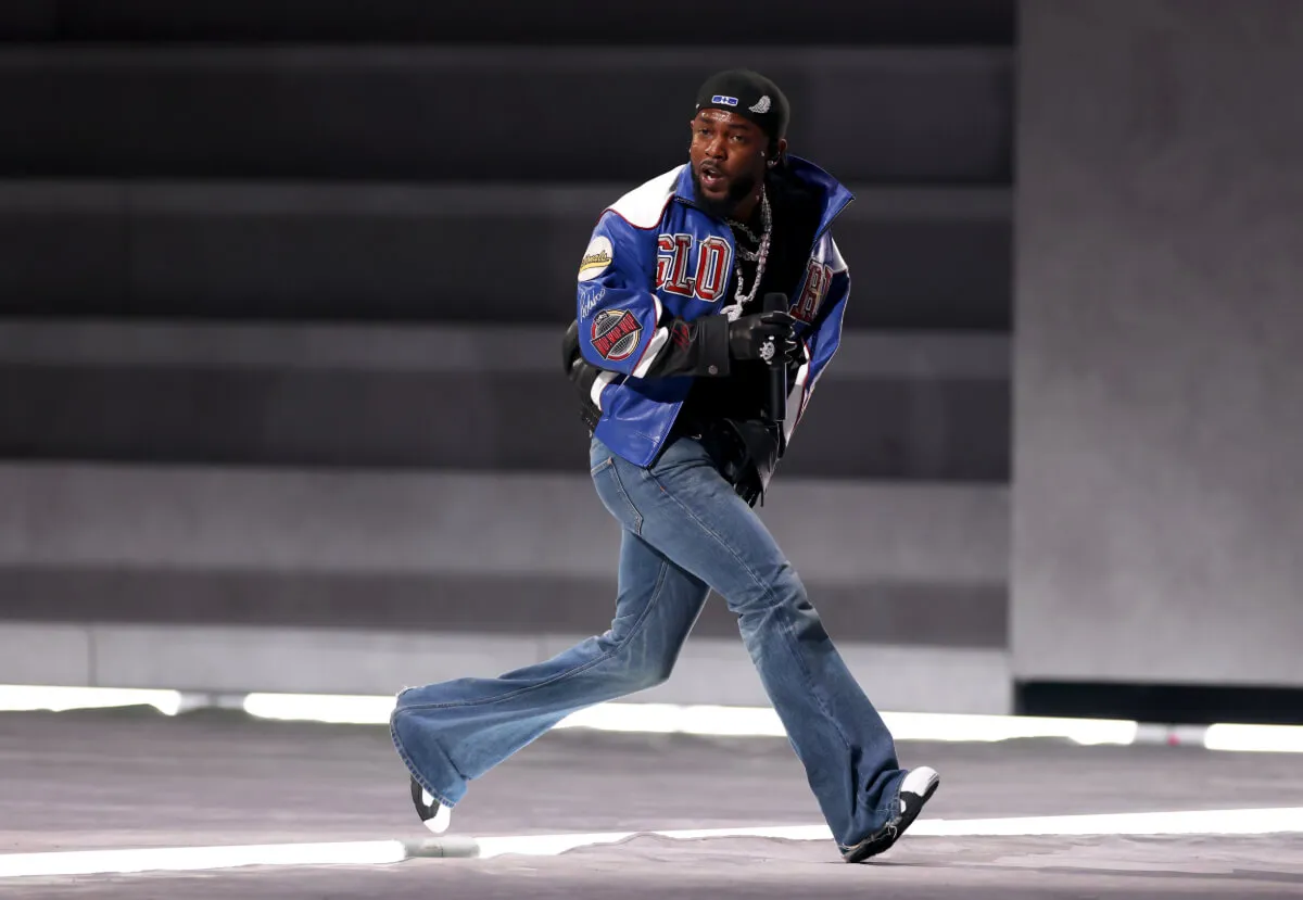 Kendrick Lamar porte un jean et une veste bleue. Il traverse une scène pendant le Super Bowl.
