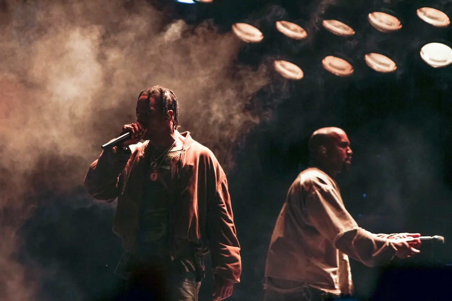 Une photo sombre et enfumée de Travis Scott jouant avec Kanye 'Ye' West en arrière-plan sur scène