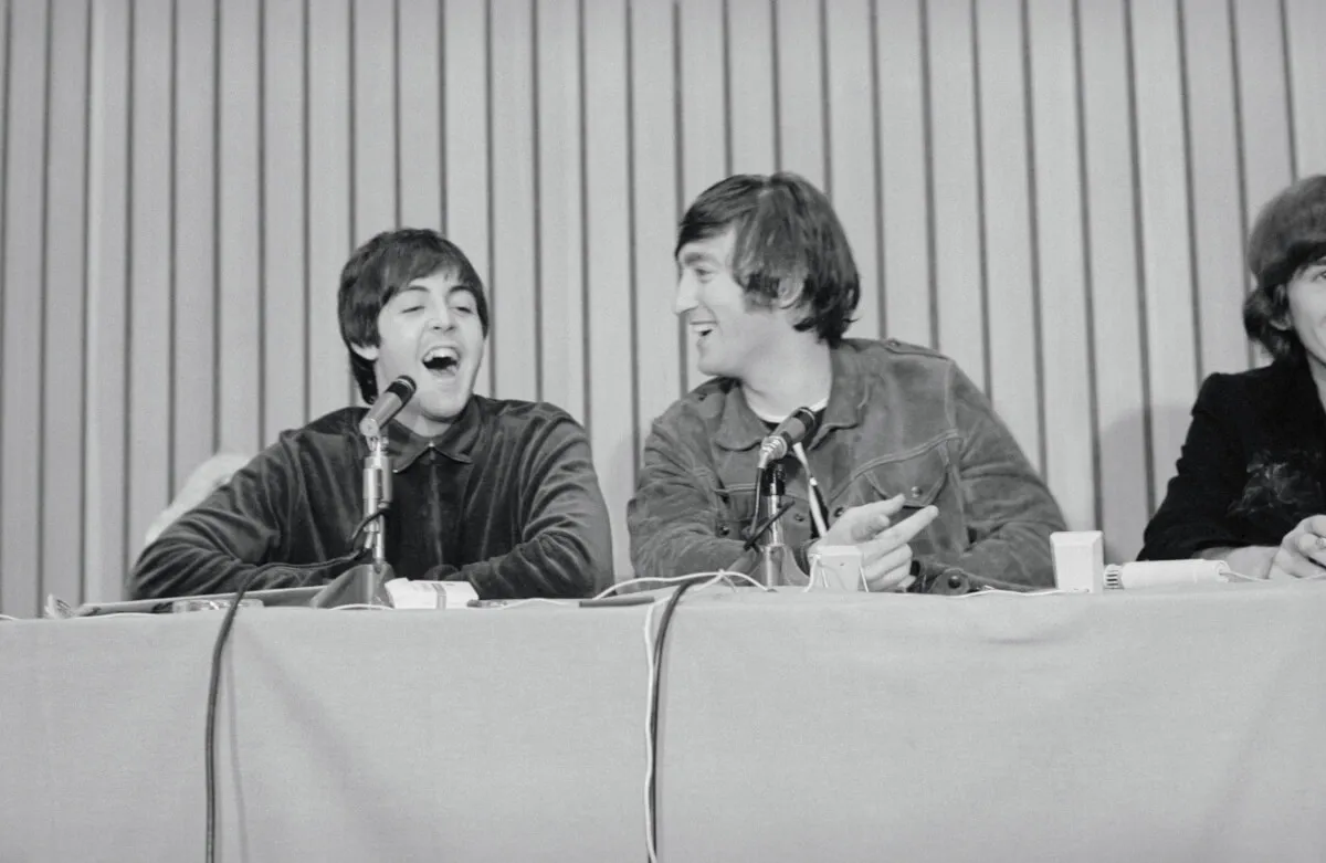 Une photo en noir et blanc de Paul McCartney et John Lennon assis devant des micros et riant.