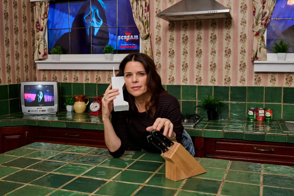 Neve Campbell se penche sur le comptoir de la cuisine de la maison « Scream ». Elle parle au téléphone et touche un bloc de couteaux. Ghost Face est dans la fenêtre derrière elle.