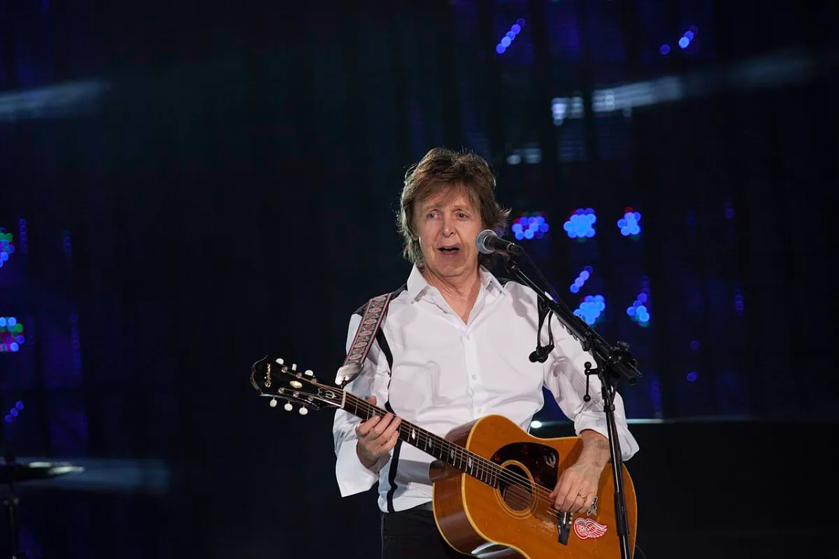 Paul McCartney porte une chemise blanche et tient une guitare acoustique. Il est debout derrière un micro.